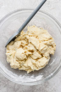 A glass bowl containing a rough, unbaked dough mixture with a spatula resting on the side.