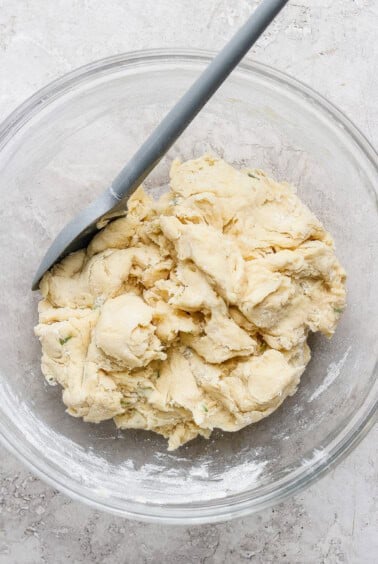 A glass bowl containing a rough, unbaked dough mixture with a spatula resting on the side.