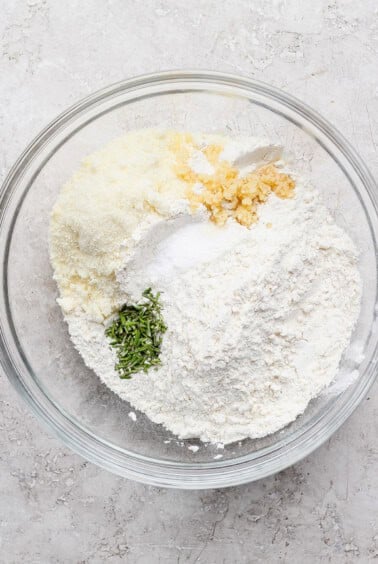 A glass bowl containing flour, grated cheese, chopped garlic, salt, and chopped fresh herbs on a light textured surface.