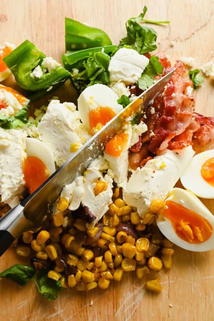 Knife slicing through soft-boiled eggs, feta cheese, cooked bacon, corn, chopped green pepper, and cilantro on a wooden cutting board.