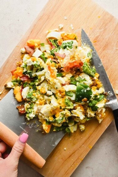 Chopped salad ingredients including hard-boiled eggs, avocado, bacon, and vegetables on a wooden cutting board with a large knife.