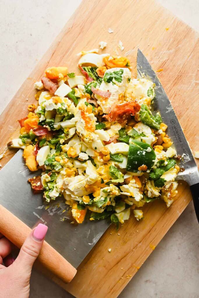 Chopped salad ingredients including hard-boiled eggs, avocado, bacon, and vegetables on a wooden cutting board with a large knife.