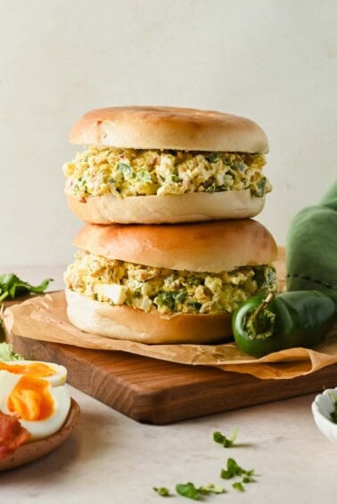 Two bagel sandwiches filled with egg salad sit stacked on a wooden board, next to a green chili pepper and sliced boiled eggs.