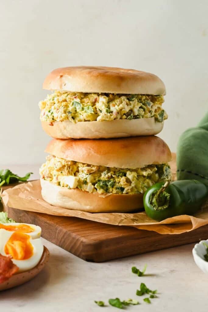 Two bagel sandwiches filled with egg salad sit stacked on a wooden board, next to a green chili pepper and sliced boiled eggs.