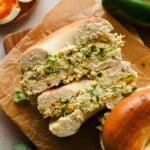 A bagel sliced in half and filled with a chunky egg salad, garnished with chopped herbs, sits on brown parchment paper.