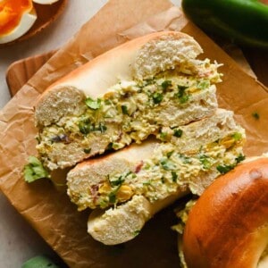 A bagel sliced in half and filled with a chunky egg salad, garnished with chopped herbs, sits on brown parchment paper.