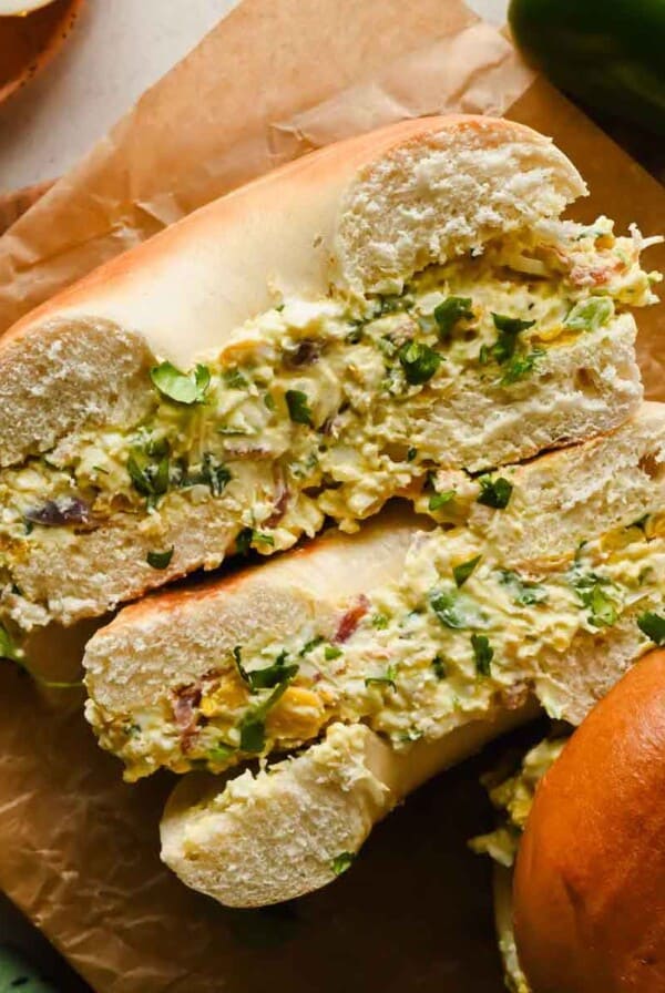 A bagel sliced in half and filled with a chunky egg salad, garnished with chopped herbs, sits on brown parchment paper.