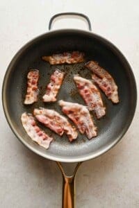 Several strips of bacon sizzling in a frying pan on a light-colored surface.