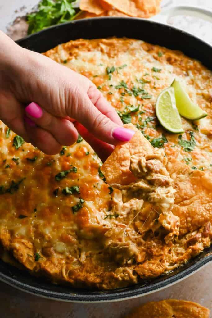A hand with pink nail polish dips a tortilla chip into a cheesy baked dip in a skillet, garnished with herbs and lime wedges.