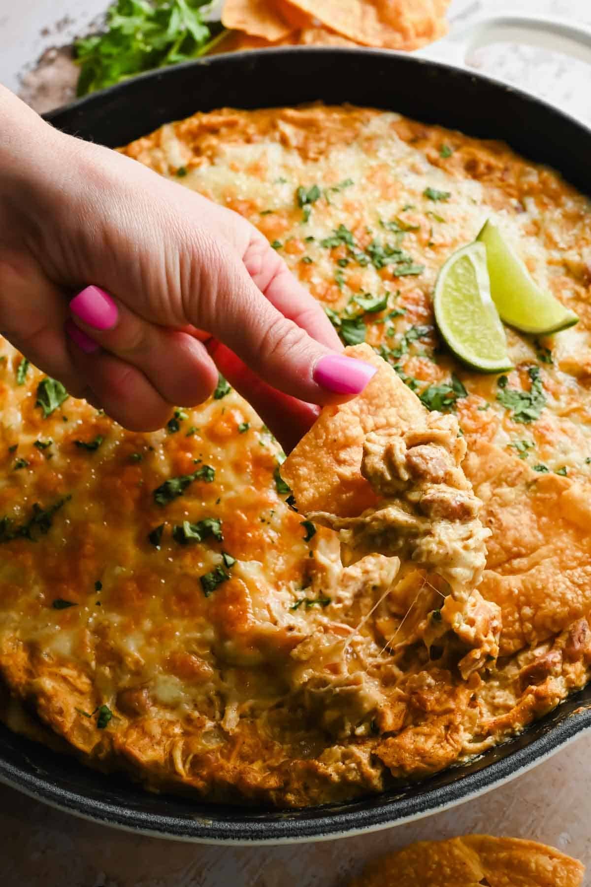 A hand with pink nails scoops cheesy dip from a skillet using a tortilla chip; the dip is topped with melted cheese, herbs, and lime wedges.
