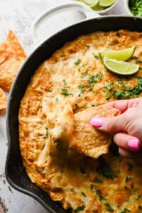 A hand with pink nail polish dips a tortilla chip into a cheesy baked dip in a skillet, garnished with fresh herbs and lime wedges.