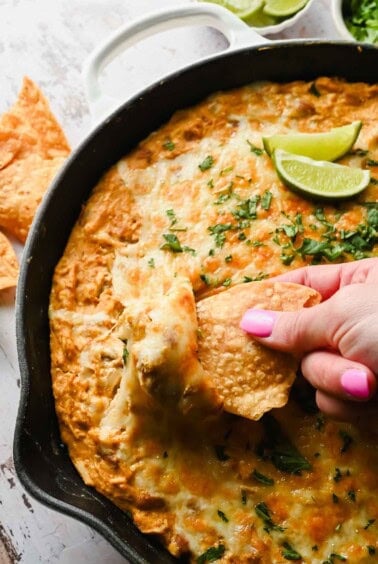 A hand with pink nail polish dips a tortilla chip into a cheesy baked dip in a skillet, garnished with fresh herbs and lime wedges.
