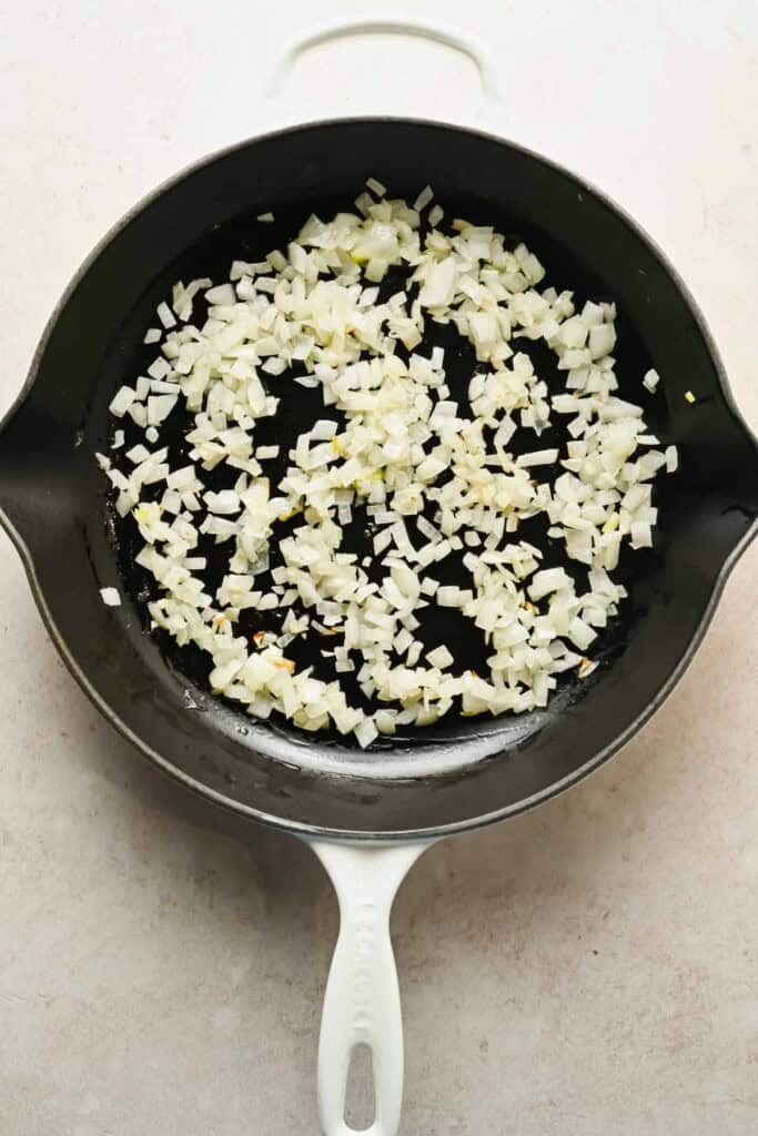 Diced onions being saut&eacute;ed in a black skillet with a white handle on a light-colored countertop.