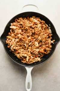 A white skillet filled with shredded chicken, beans, and spices on a light-colored surface.