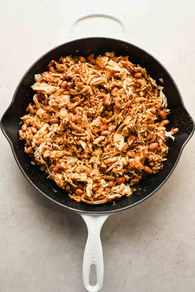 A white skillet filled with shredded chicken, beans, and spices on a light-colored surface.