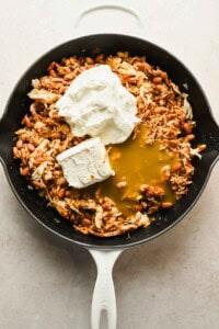 A cast iron skillet containing shredded chicken, pinto beans, cream cheese, sour cream, and green enchilada sauce before mixing.