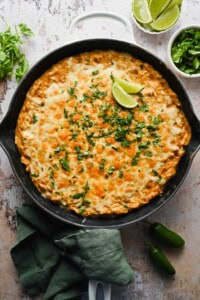 A baked casserole topped with melted cheese and chopped cilantro, garnished with lime wedges in a black skillet, surrounded by fresh herbs and jalapeños.