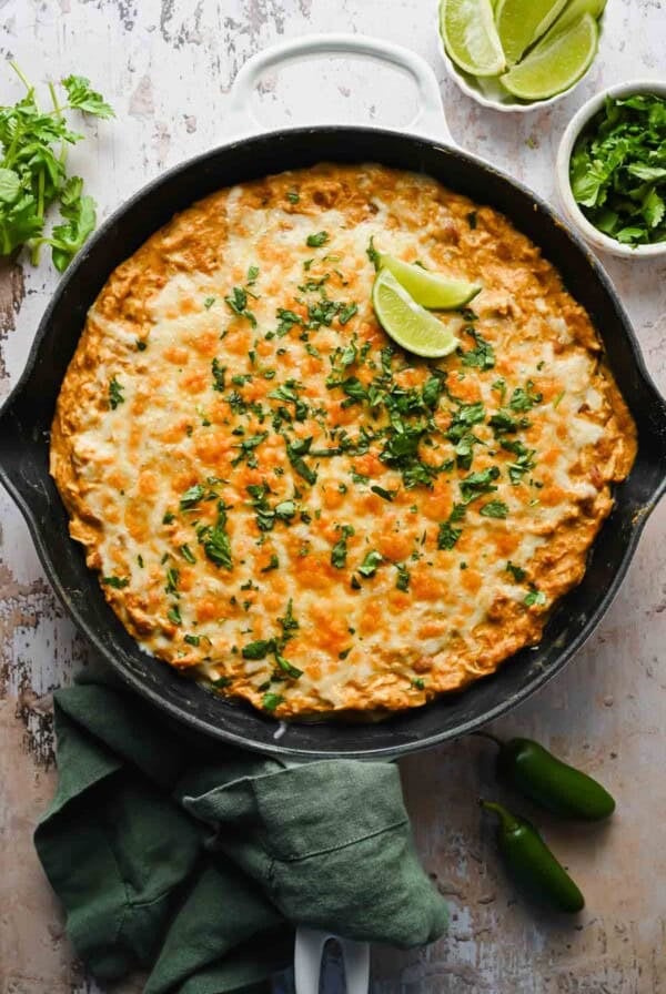 A baked casserole topped with melted cheese and chopped cilantro, garnished with lime wedges in a black skillet, surrounded by fresh herbs and jalapeños.