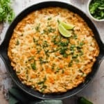 A skillet filled with cheesy baked casserole topped with chopped cilantro and two lime wedges, set on a rustic white surface.