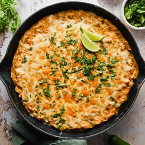 A skillet filled with cheesy baked casserole topped with chopped cilantro and two lime wedges, set on a rustic white surface.
