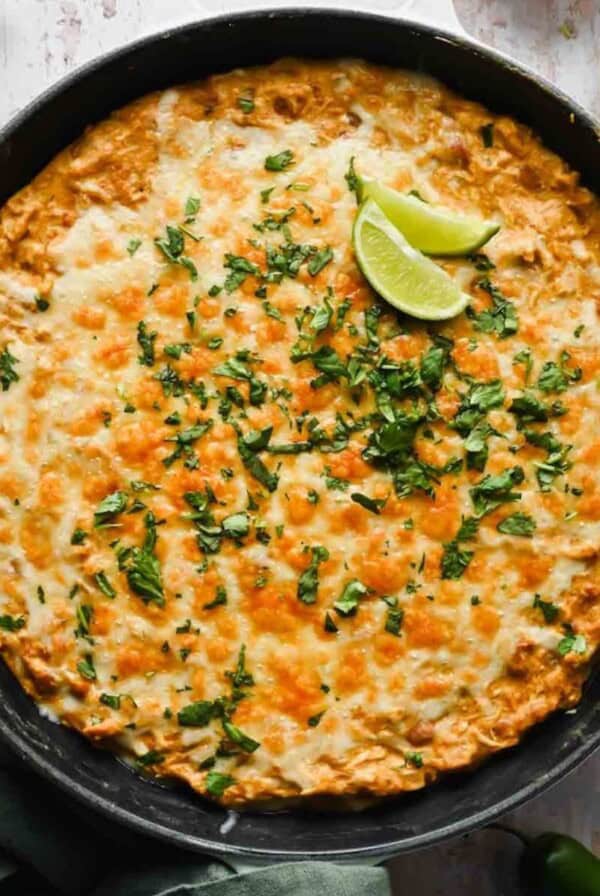 A skillet filled with cheesy baked casserole topped with chopped cilantro and two lime wedges, set on a rustic white surface.