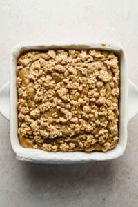 A square baking dish filled with baked crumb-topped coffee cake, photographed from above on a light surface.