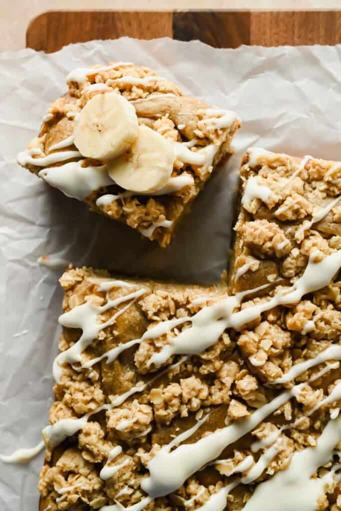 A square piece of banana coffee cake with oat crumble topping and white icing, garnished with banana slices, sits next to the main cake on parchment paper.