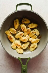 Sliced bananas being sautéed in a gray pan with a green handle on a light-colored surface.