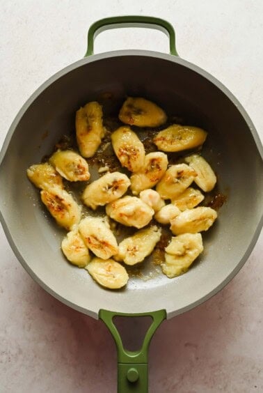 Sliced bananas being saut&eacute;ed in a gray pan with a green handle on a light-colored surface.