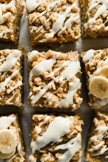 Squares of baked oatmeal topped with white icing drizzle and oat crumble, with some pieces garnished with banana slices.