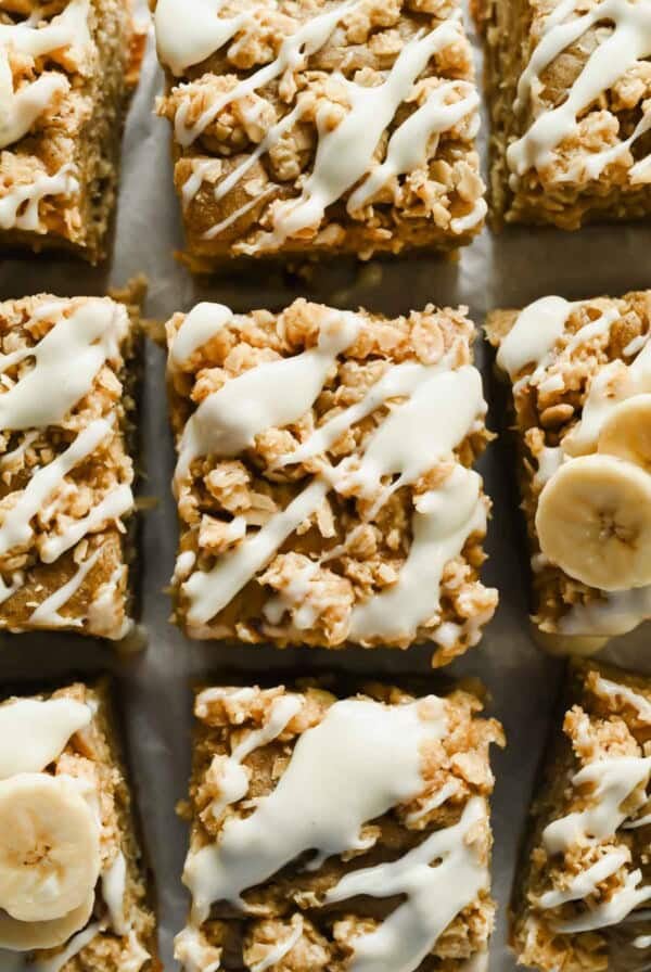 Squares of baked oatmeal topped with white icing drizzle and oat crumble, with some pieces garnished with banana slices.