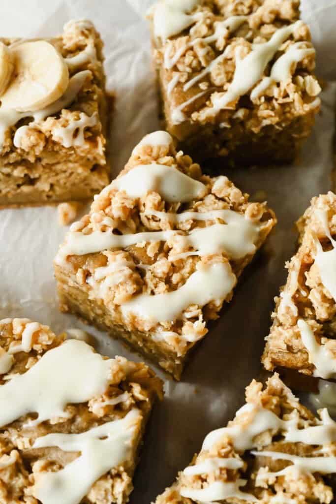 Close-up of square banana oat bars topped with a white icing drizzle, arranged on parchment paper.