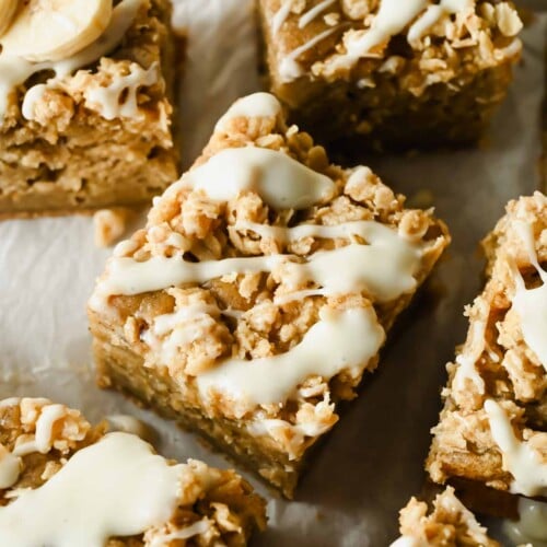 Close-up of square banana oat bars topped with a white icing drizzle, arranged on parchment paper.
