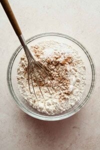 A whisk rests in a glass bowl containing a mixture of flour and brown spices on a light countertop.