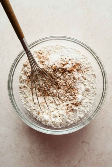 A whisk rests in a glass bowl containing a mixture of flour and brown spices on a light countertop.