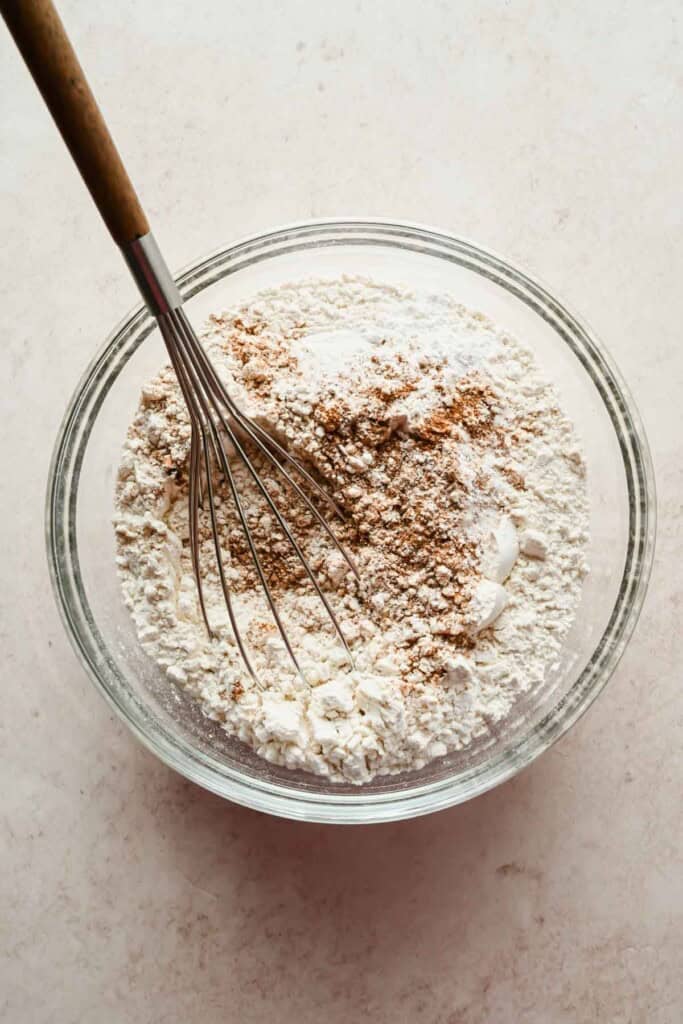 A whisk rests in a glass bowl containing a mixture of flour and brown spices on a light countertop.