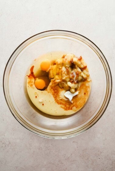 A glass mixing bowl containing eggs, mashed banana, yogurt, and vanilla extract on a light countertop.