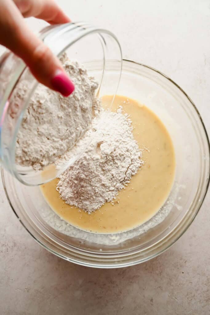 A hand pours flour from a glass bowl into a larger bowl containing a beige batter on a light countertop.