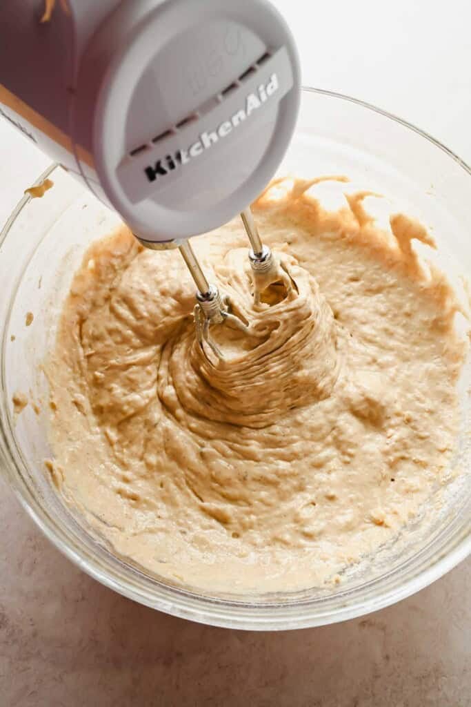 A KitchenAid hand mixer blends thick batter in a clear glass bowl on a light-colored surface.