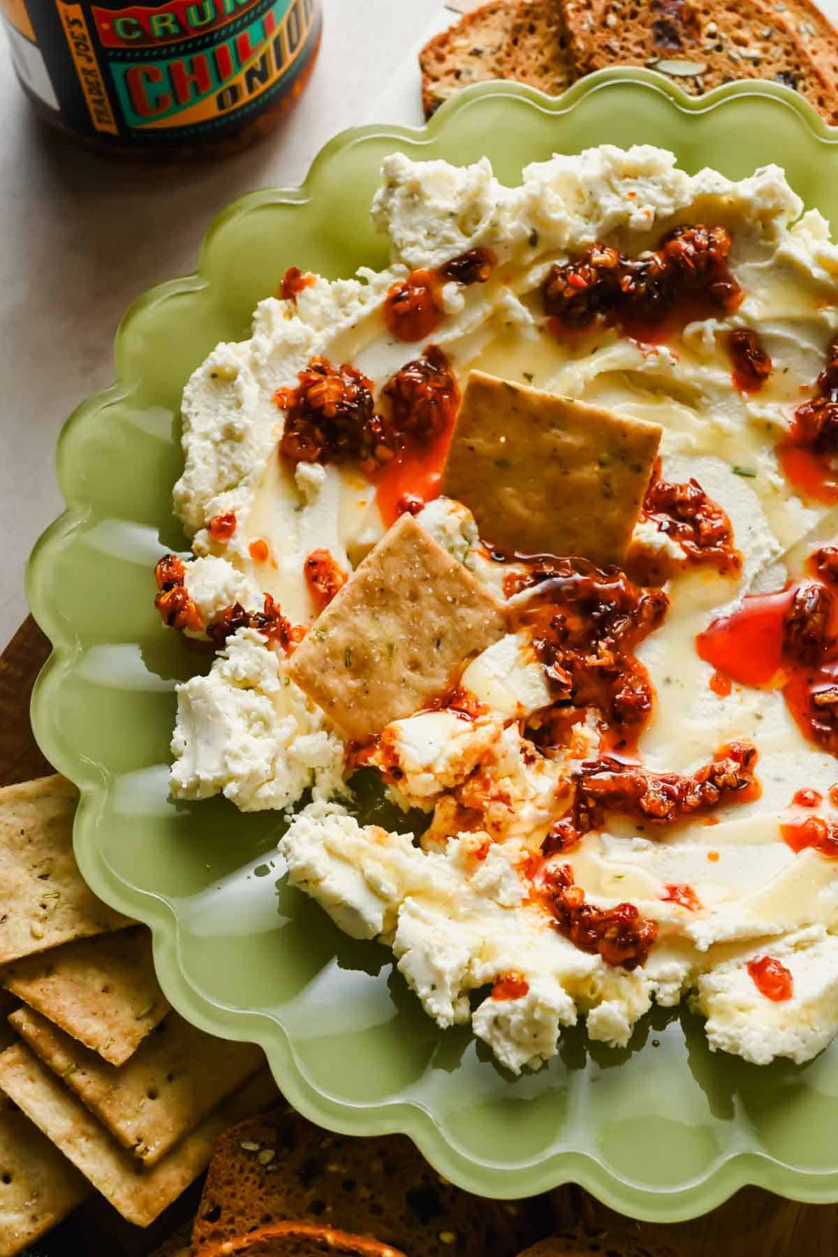 Boursin cheese topped with chili crunch oil and honey in a green bowl with crackers