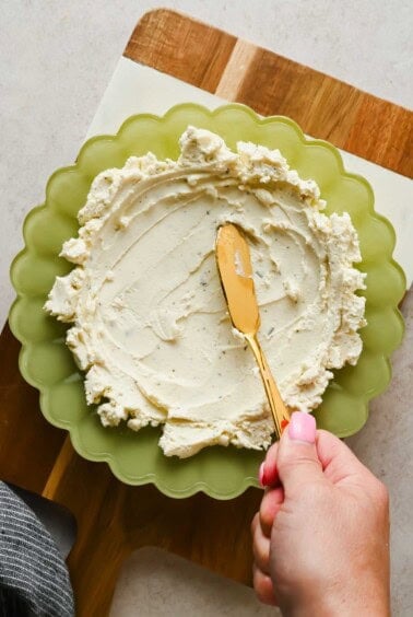 Spreading Boursin cheese into a green scalloped bowl with a gold knife