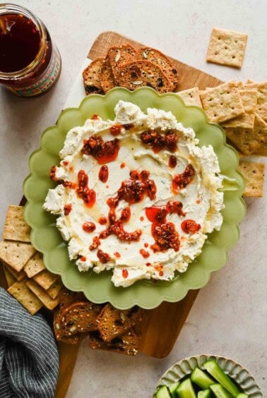Boursin chili crunch dip on a board with crackers, toast, cucumber slices, and a jar of chili crunch