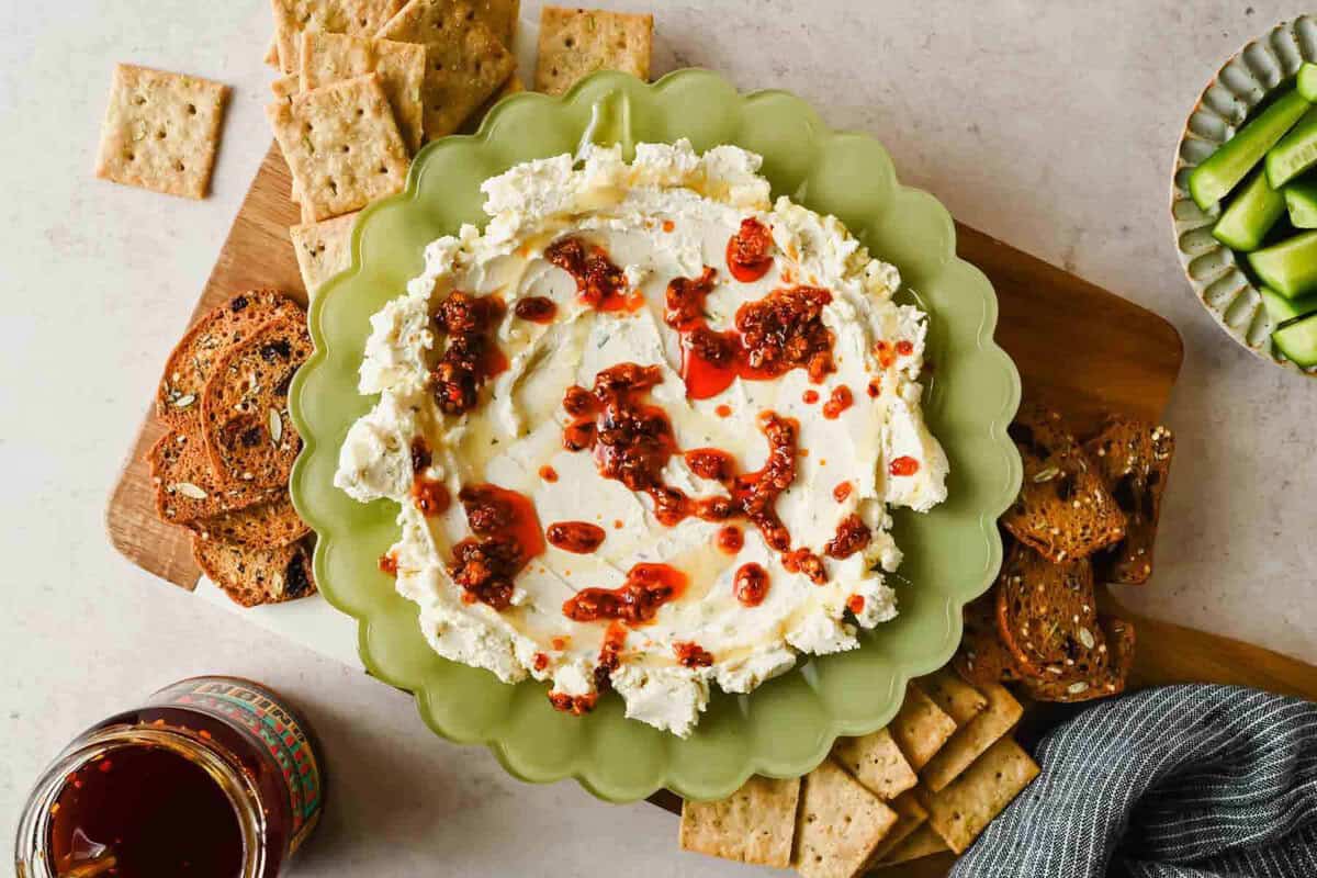 Boursin chili crunch dip on a board with crackers, toast, cucumber slices, and a jar of chili crunch