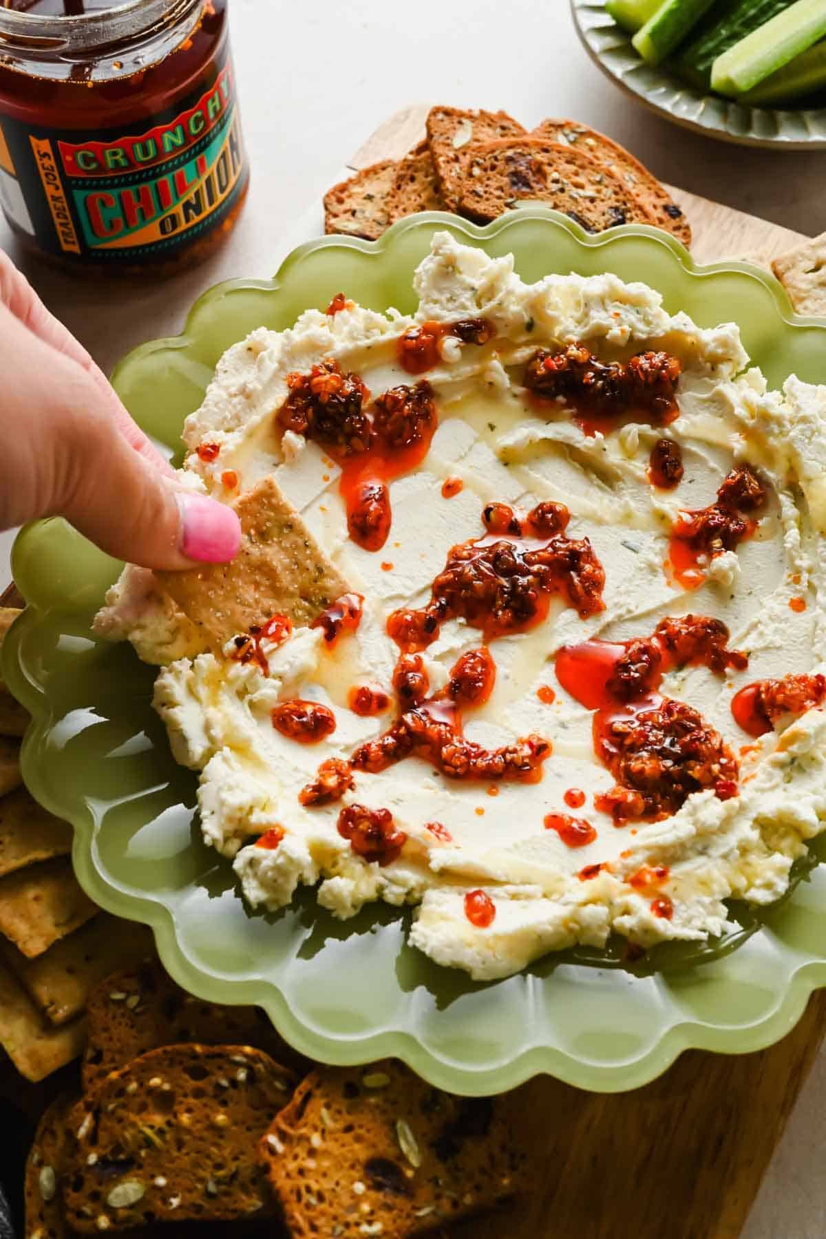 A hand dipping a cracker into boursin chili crunch dip surrounded by crackers and vegetables