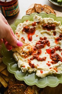 A hand dipping a cracker into whipped cheese topped with chili oil, served on a green plate with more crackers on the side.