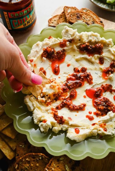 A hand dipping a cracker into whipped cheese topped with chili oil, served on a green plate with more crackers on the side.