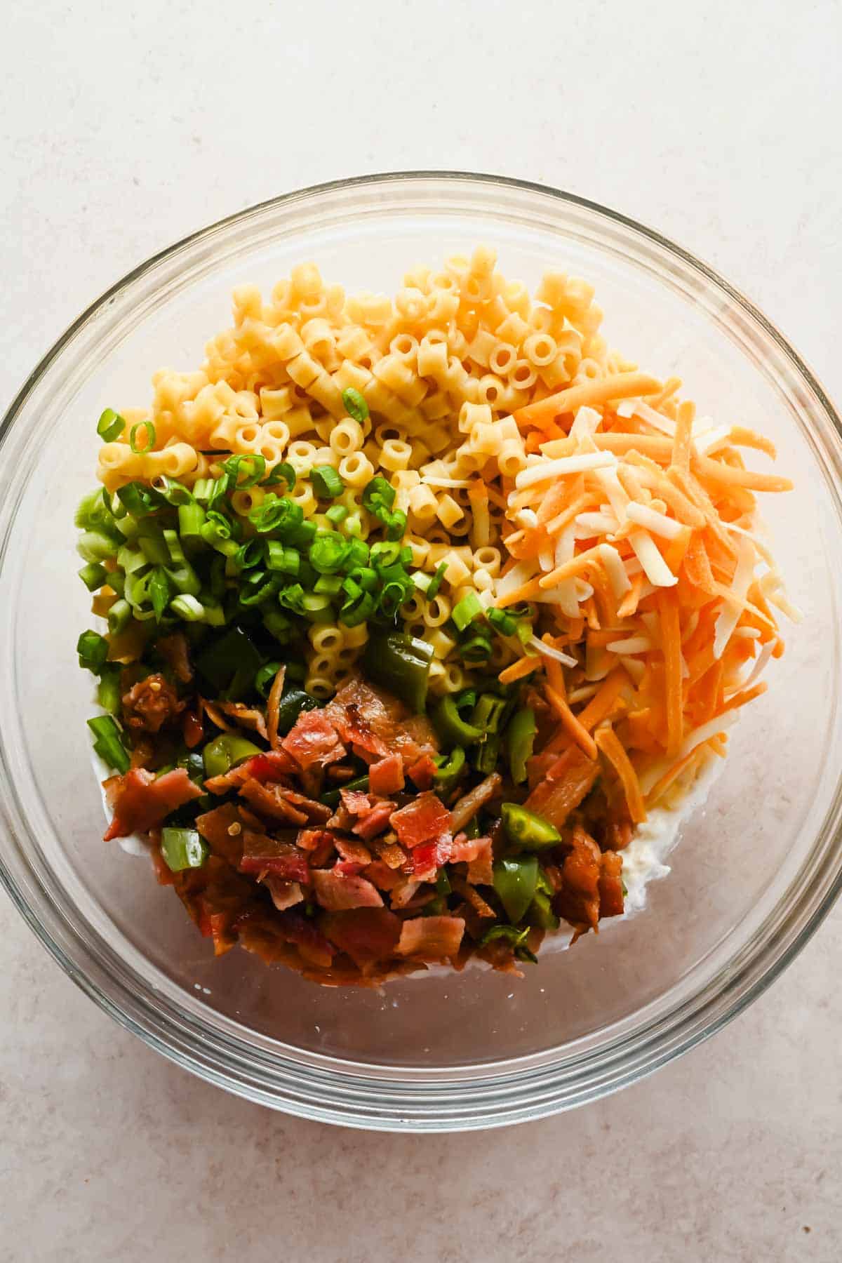 Glass bowl with ditalini pasta, shredded cheddar, chopped green onions, crispy bacon, and roasted jalape&ntilde;os ready to toss