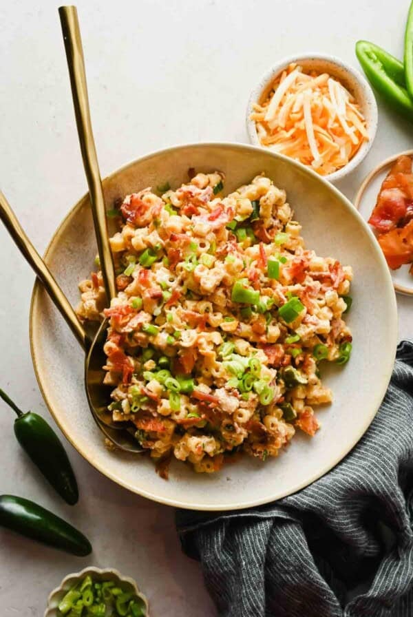 Jalape&ntilde;o popper pasta salad in a large bowl with crispy bacon, green onions, and shredded cheese, with gold utensils and fresh jalape&ntilde;os nearby