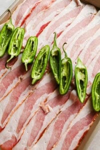 Sliced bacon and halved jalapeño peppers on a tray before roasting