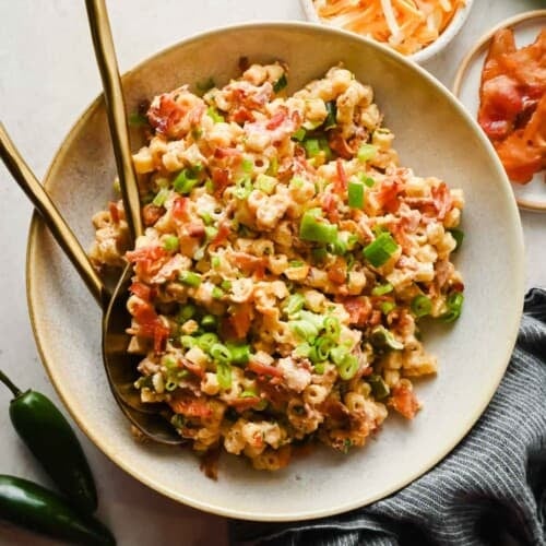 Jalape&ntilde;o popper pasta salad topped with green onions and bacon in a bowl with serving utensils